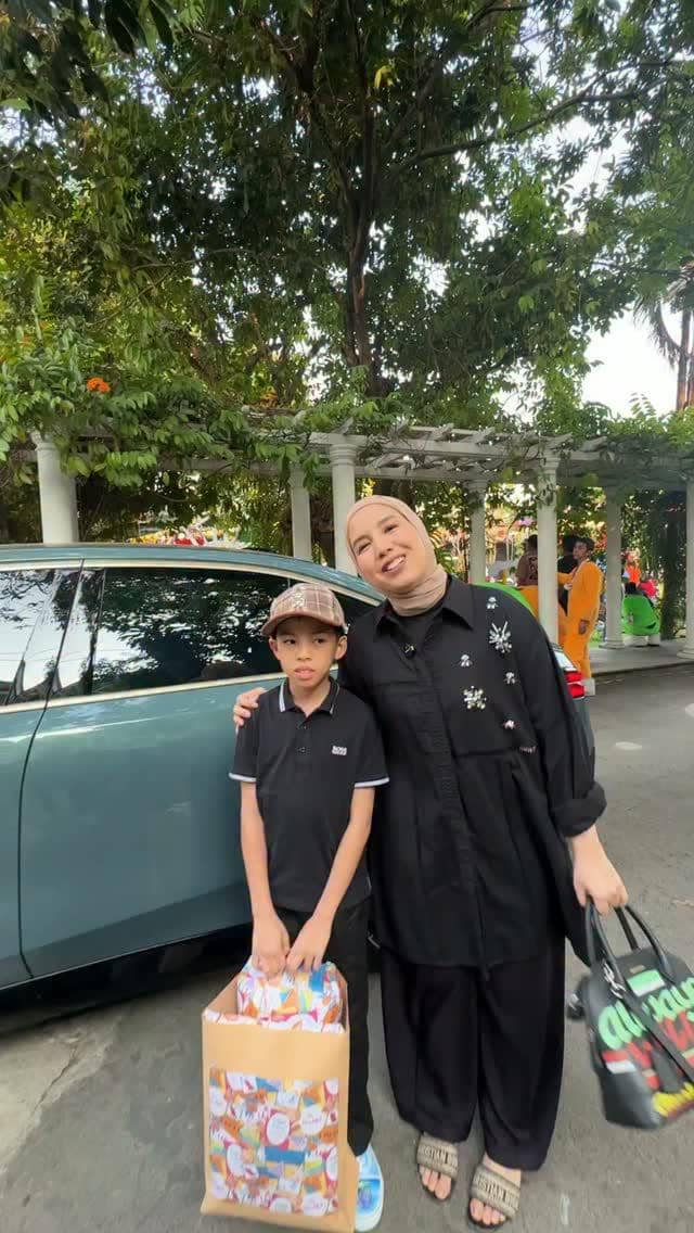 Seorang wanita berbusana hitam berpose bersama anak laki-laki yang membawa tas kado besar di samping mobil berwarna biru kehijauan.