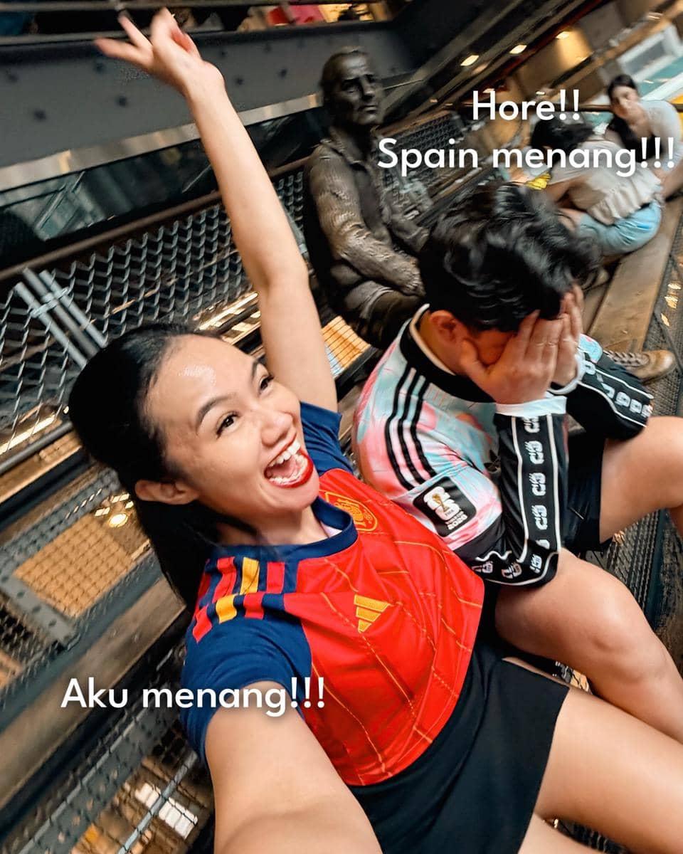 Seorang wanita mengenakan jersey tim Spanyol bersorak gembira sambil berfoto selfie dengan seorang pria yang menutupi wajahnya setelah pertandingan Piala Dunia 2026.