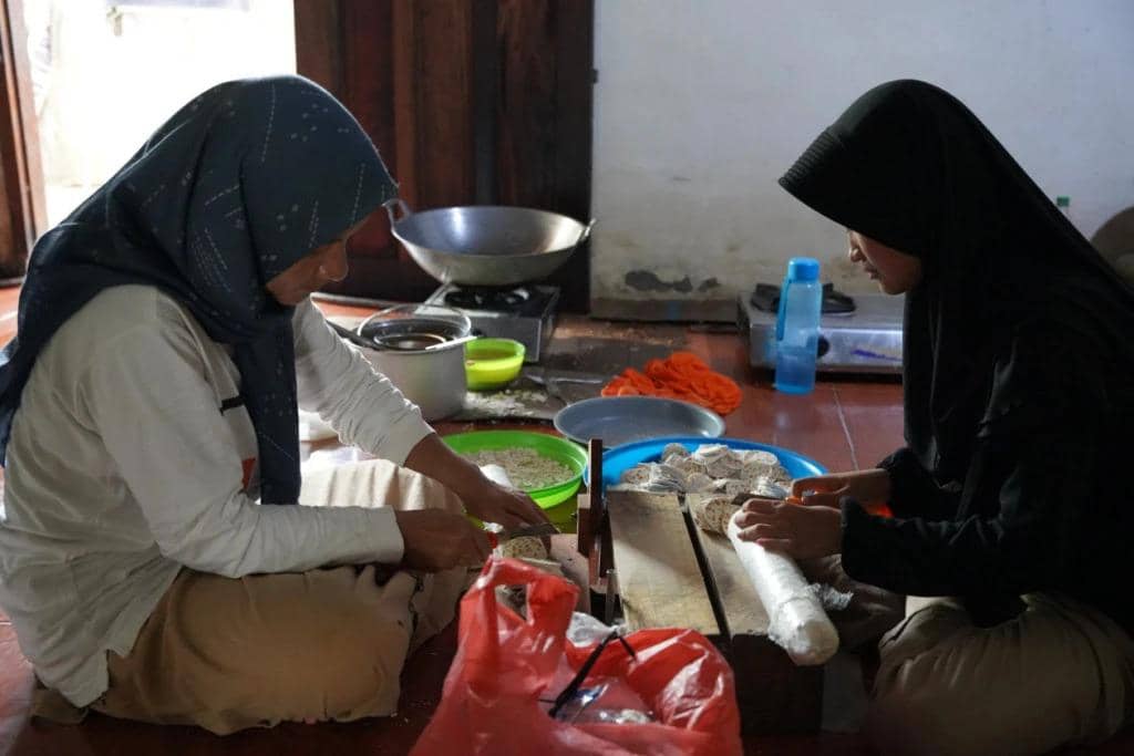 Dua perempuan berhijab duduk di lantai dapur sederhana sambil menyiapkan makanan dengan peralatan tradisional di depan mereka.