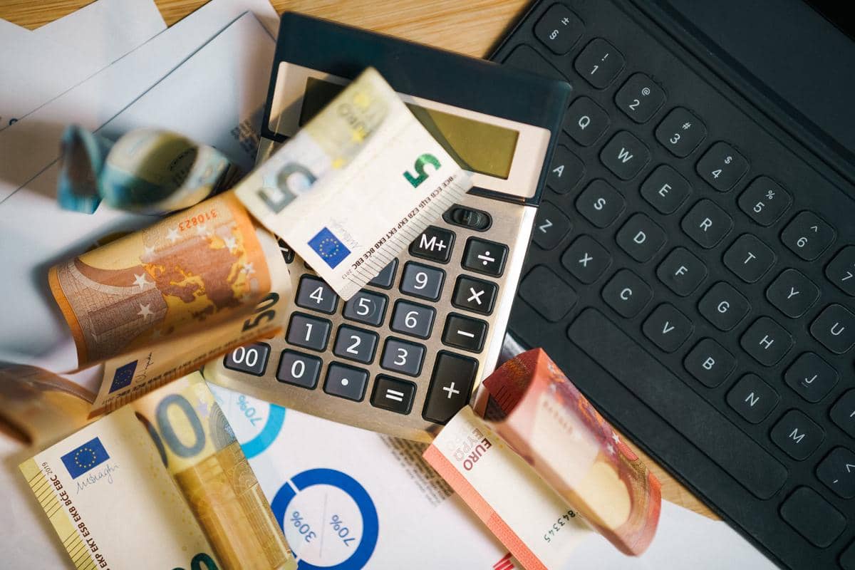 Uang euro, kalkulator, dan keyboard di atas meja dengan lembar grafik keuangan menggambarkan analisis risiko keuangan.