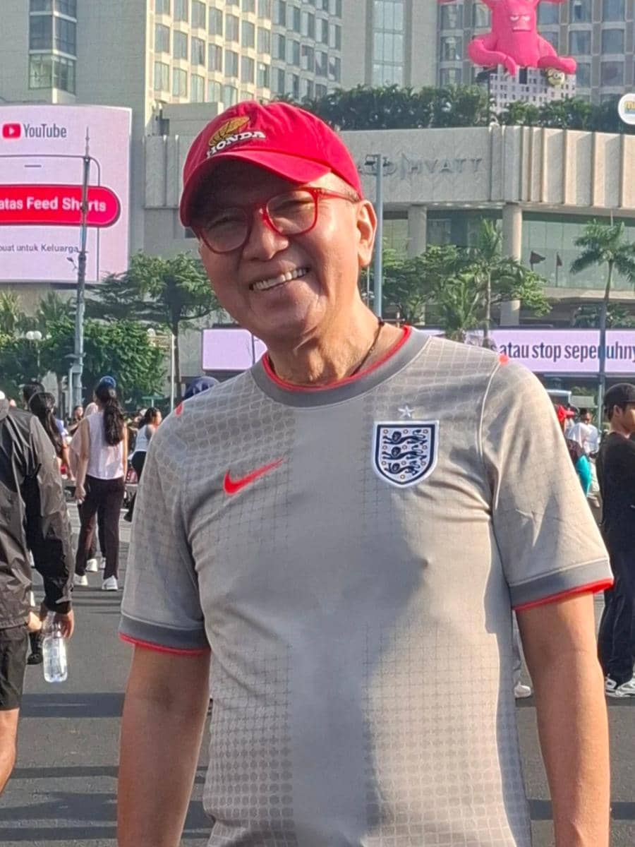 Seorang pria mengenakan kaus tim nasional Inggris dan topi merah tersenyum di area luar ruangan dengan latar gedung bertingkat.