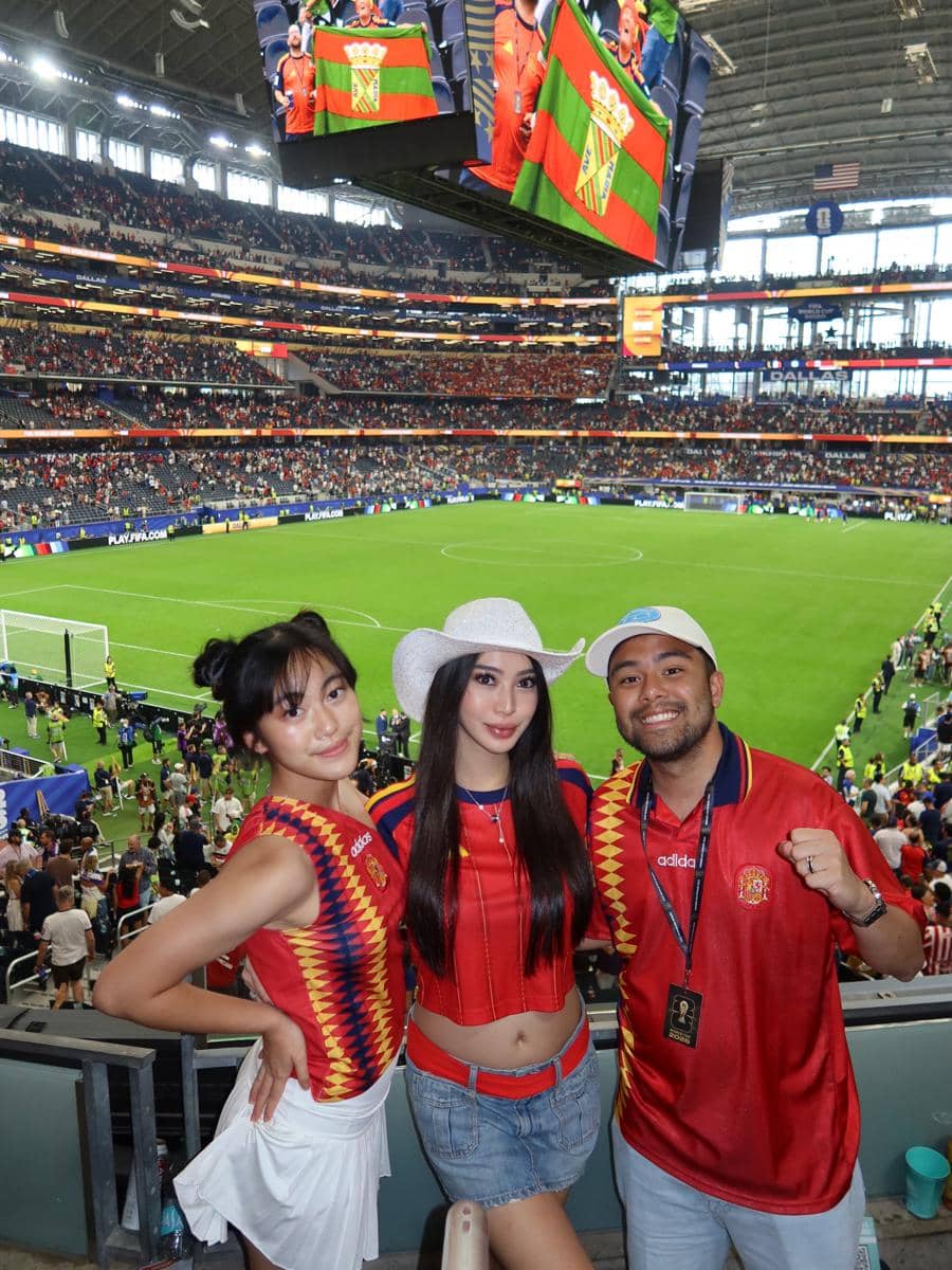 Tiga orang mengenakan pakaian berwarna merah dan kuning berpose di tribun stadion besar saat pertandingan sepak bola Spanyol versus Prancis berlangsung.