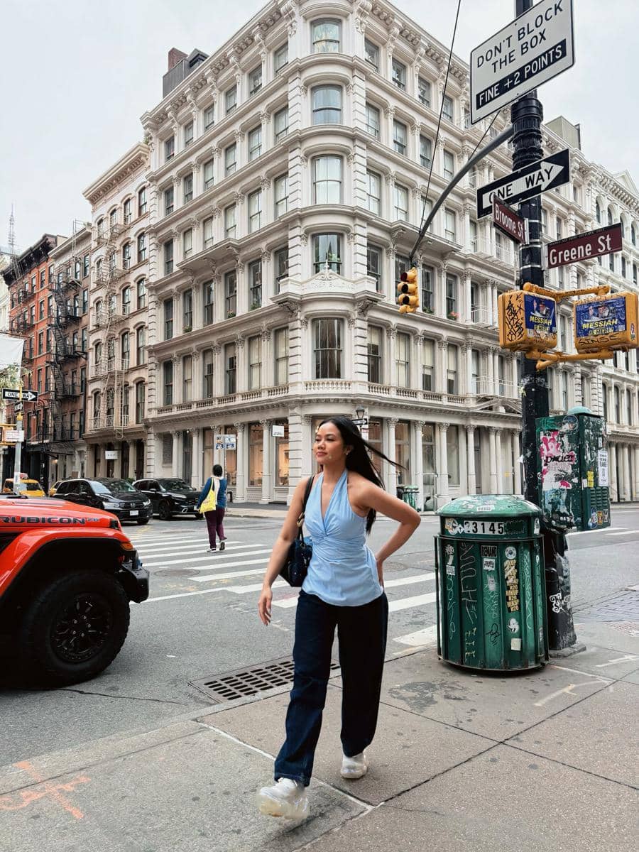 Seorang wanita berjalan di persimpangan jalan kawasan SoHo, New York, dengan latar gedung bergaya klasik dan suasana kota yang ramai.