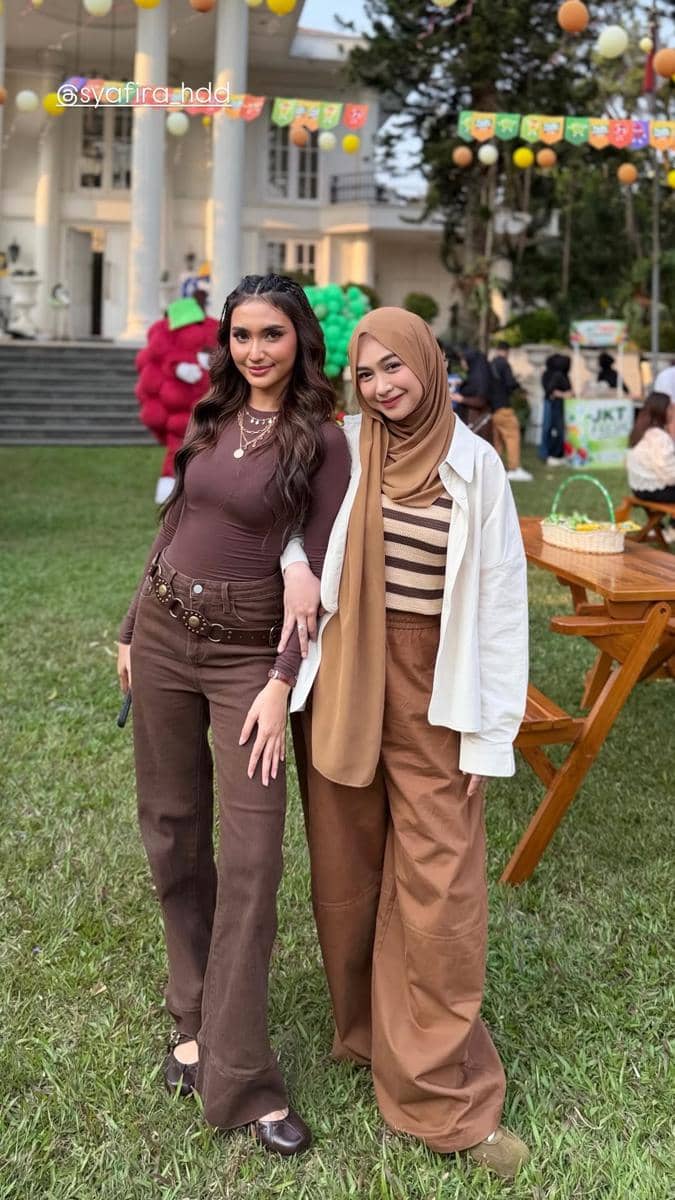 Dua wanita berpose bersama di taman acara ulang tahun dengan dekorasi balon warna-warni dan bangunan besar di latar belakang.