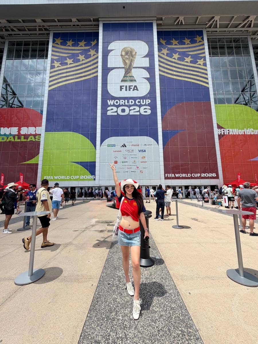 Seorang perempuan mengenakan jersey merah dan topi putih berpose gembira di depan stadion dengan spanduk besar FIFA World Cup 2026.