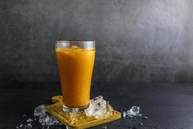 Segelas jamu kunyit asam berwarna oranye cerah disajikan di atas alas kayu dengan es batu di sekitarnya di permukaan gelap.