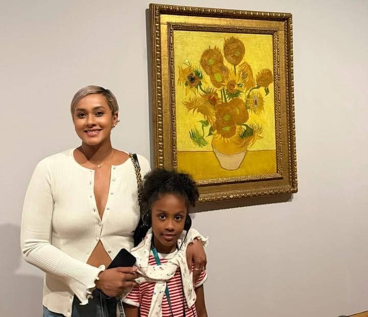 Seorang wanita dan anak perempuan berpose di depan lukisan bunga matahari terkenal di Museum Van Gogh dengan latar dinding putih.