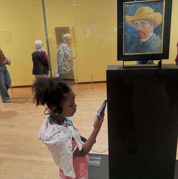 Seorang anak kecil mengenakan headphone sambil melihat layar ponsel di depan lukisan potret di Museum Van Gogh.