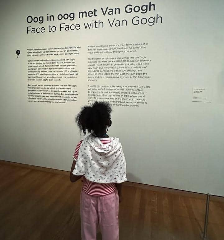 Seorang anak mengenakan pakaian merah muda berdiri di depan dinding bertulisan 'Face to Face with Van Gogh' di Museum Van Gogh Amsterdam.