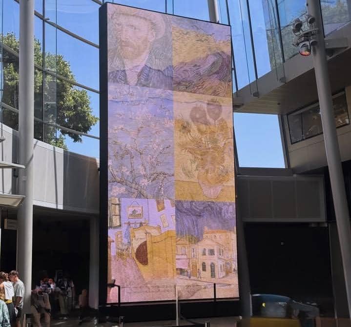 Layar digital besar menampilkan kolase karya seni Vincent van Gogh di area dalam Museum Van Gogh dengan pengunjung berjalan di sekitarnya.