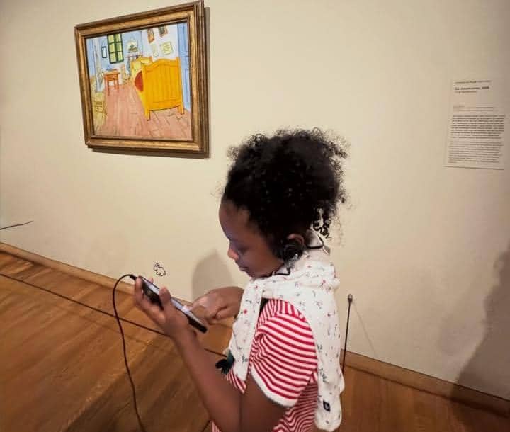 Seorang anak mengenakan headphone dan melihat ponsel di depan lukisan terkenal di Museum Van Gogh dengan suasana galeri seni.