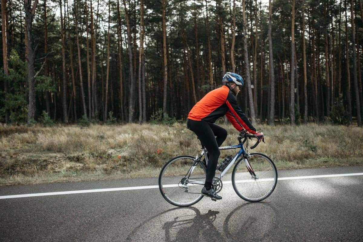 Seorang pesepeda mengenakan jaket oranye dan helm biru bersepeda di jalan aspal dengan latar belakang hutan pinus.