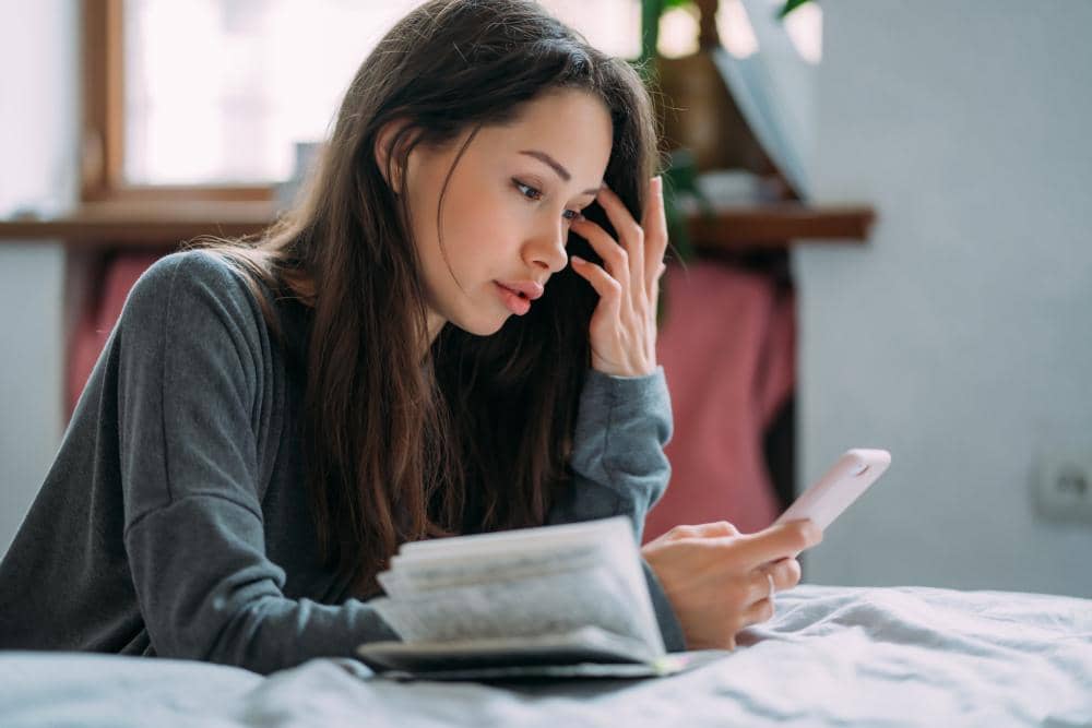 Seorang perempuan duduk sambil memegang ponsel dan membuka buku di depannya, tampak fokus melihat layar ponsel di dalam ruangan.