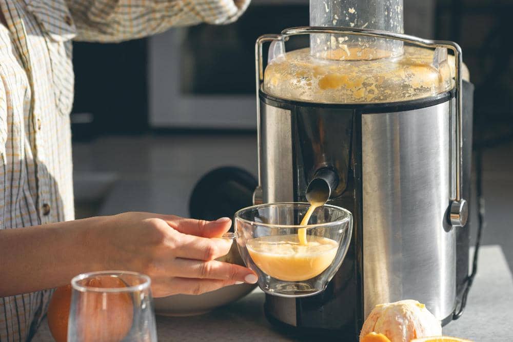 Seseorang menuangkan jus jeruk segar dari mesin blender ke dalam gelas kaca di dapur dengan pencahayaan alami.