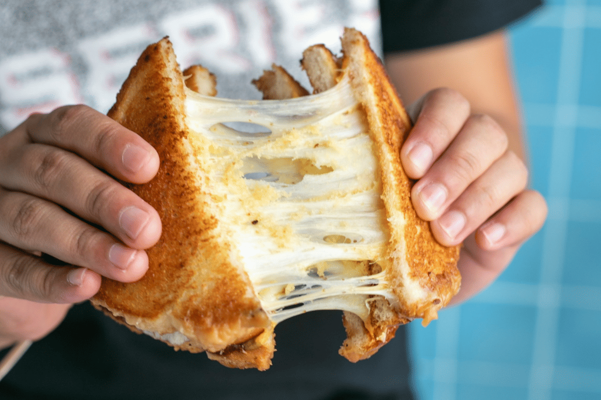 Tangan memegang roti panggang keju leleh yang ditarik hingga keju meregang di antara dua potong roti berwarna keemasan.