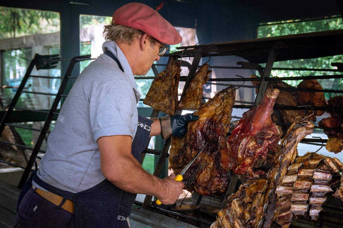 Seorang pria mengenakan topi merah dan celemek sedang memanggang potongan besar daging sapi di atas panggangan untuk asado tradisional.