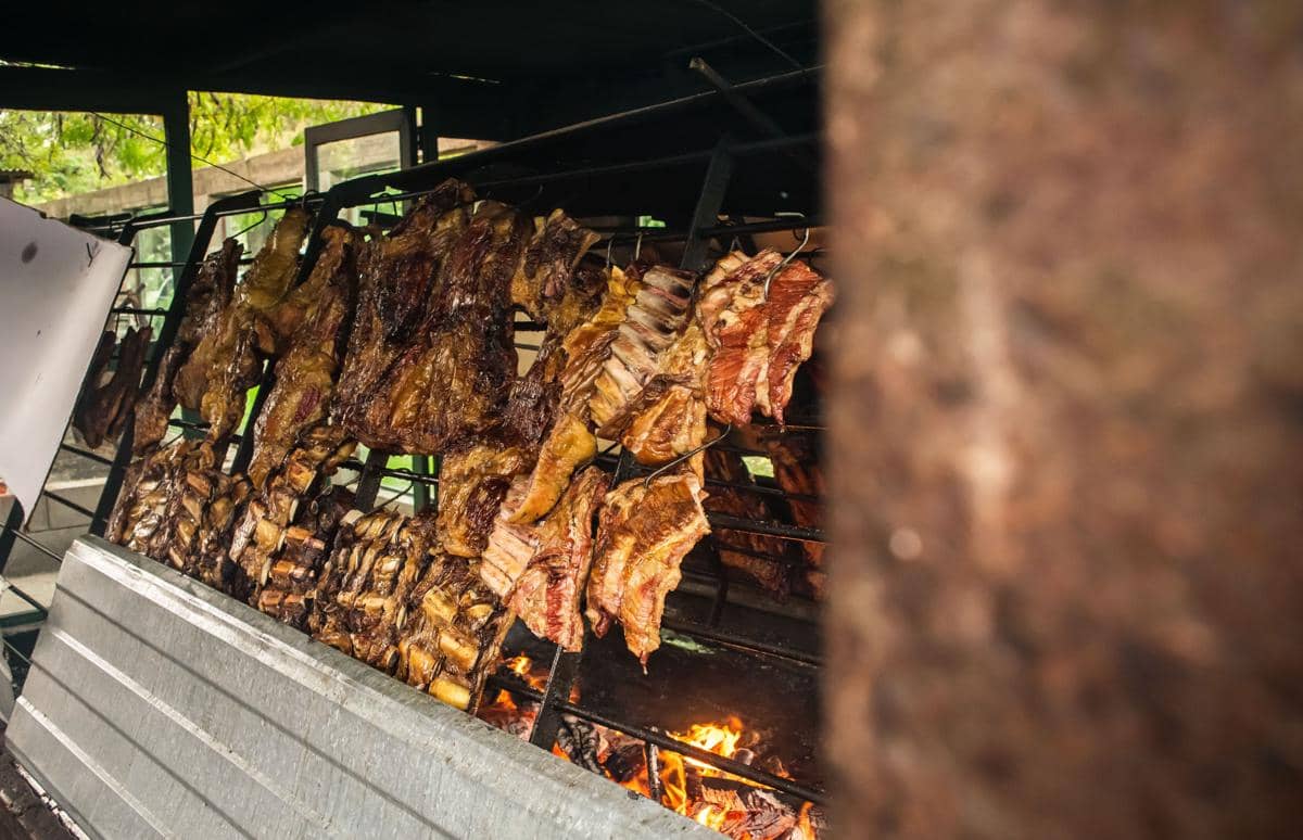 Potongan daging sapi besar dipanggang di atas bara api dengan metode asado tradisional di atas panggangan logam terbuka.