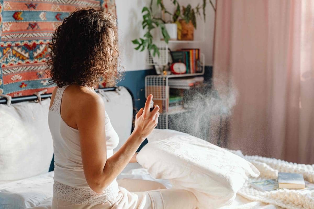 Seorang wanita menyemprotkan pengharum ruangan pada bantal di kamar tidur yang terang dengan dekorasi warna hangat dan tanaman hias.