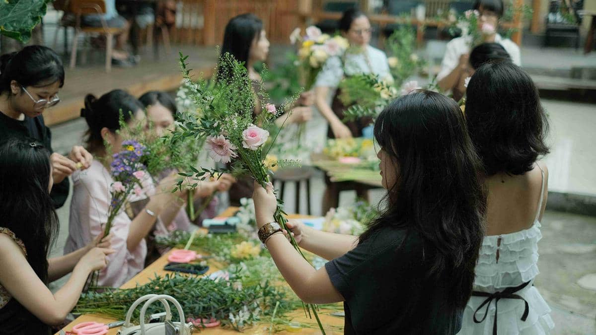 Sekelompok orang sedang mengikuti workshop merangkai bunga, menyusun berbagai jenis bunga dan daun di atas meja kayu.