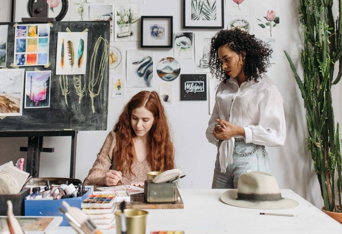 Dua perempuan sedang mengikuti workshop melukis di studio seni dengan dinding penuh karya lukisan dan alat cat air di meja.