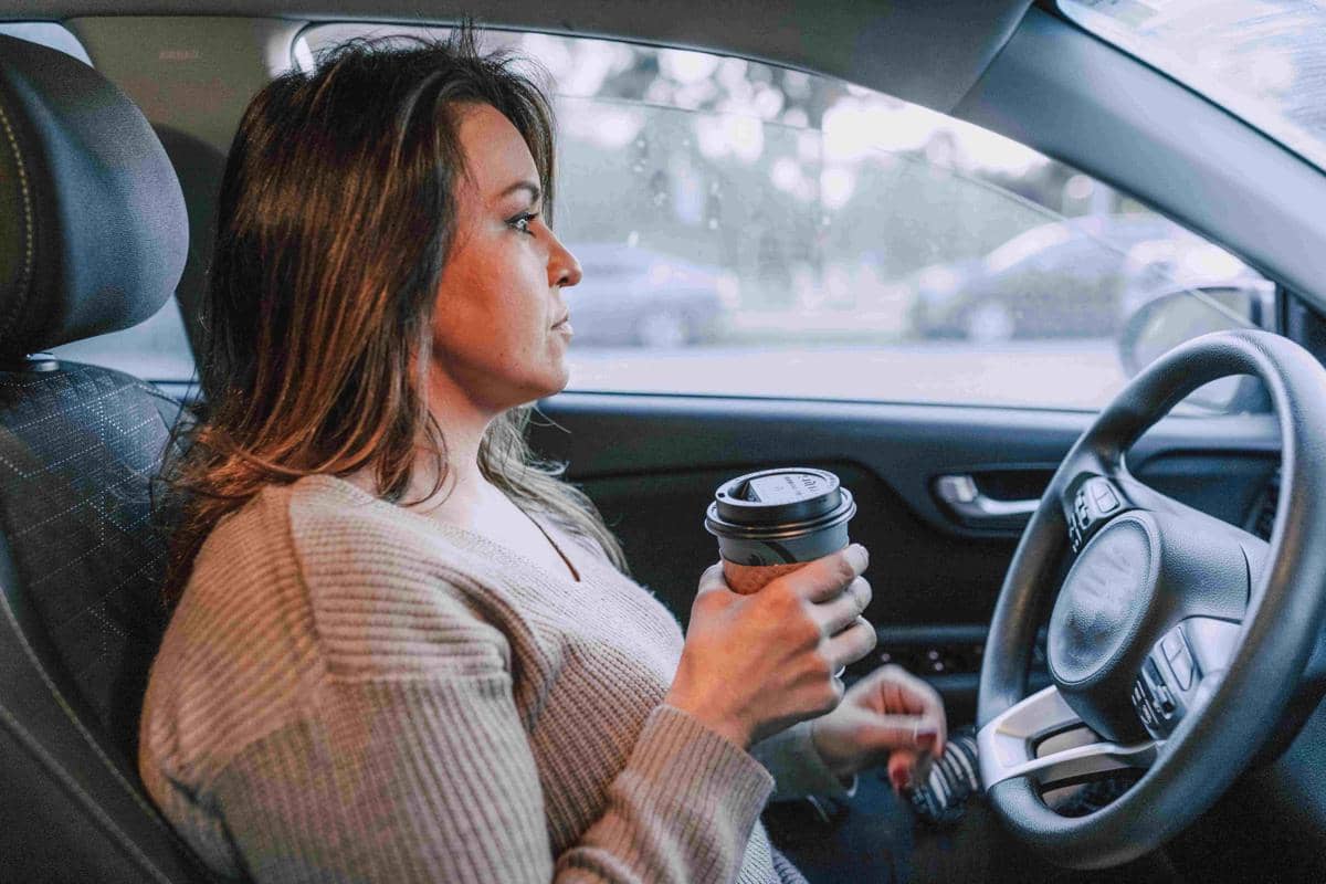 Seorang wanita duduk di kursi pengemudi mobil sambil memegang secangkir kopi dan bersiap untuk berkendara di pagi hari.