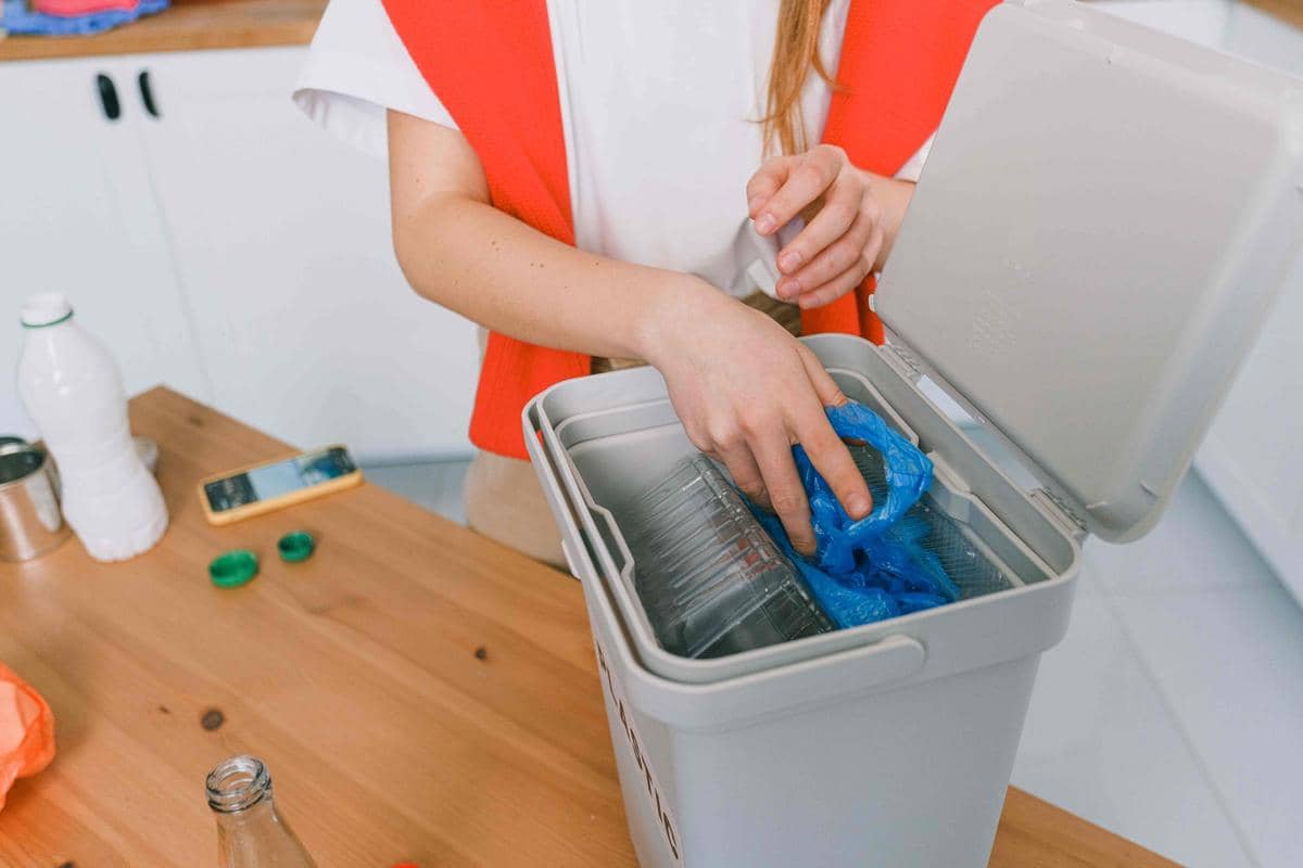 Seseorang mengenakan pakaian merah putih sedang membuang sampah plastik ke dalam tempat sampah di dapur.