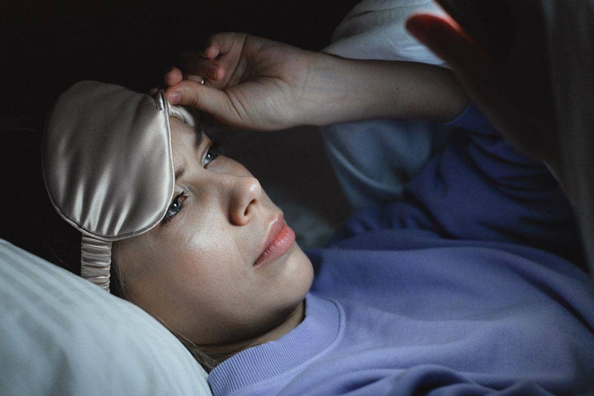 Seorang wanita mengenakan penutup mata tidur berbaring di tempat tidur sambil menatap layar ponsel di malam hari.