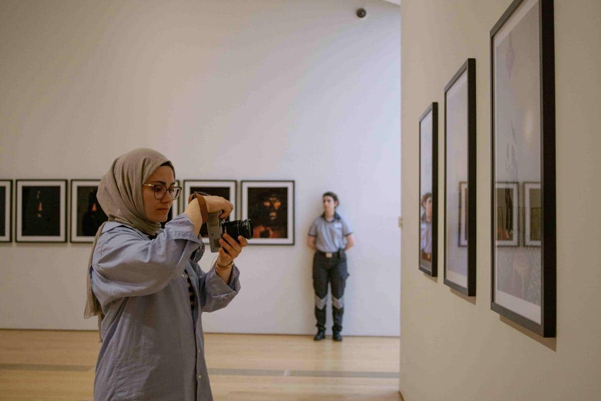 Seorang perempuan berhijab memegang kamera di galeri seni dengan beberapa foto berbingkai di dinding dan petugas berdiri di latar belakang.