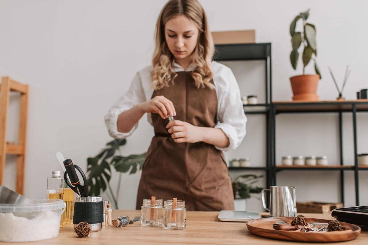 Seorang perempuan mengenakan celemek cokelat sedang menyiapkan bahan untuk membuat lilin aromaterapi di atas meja kayu.