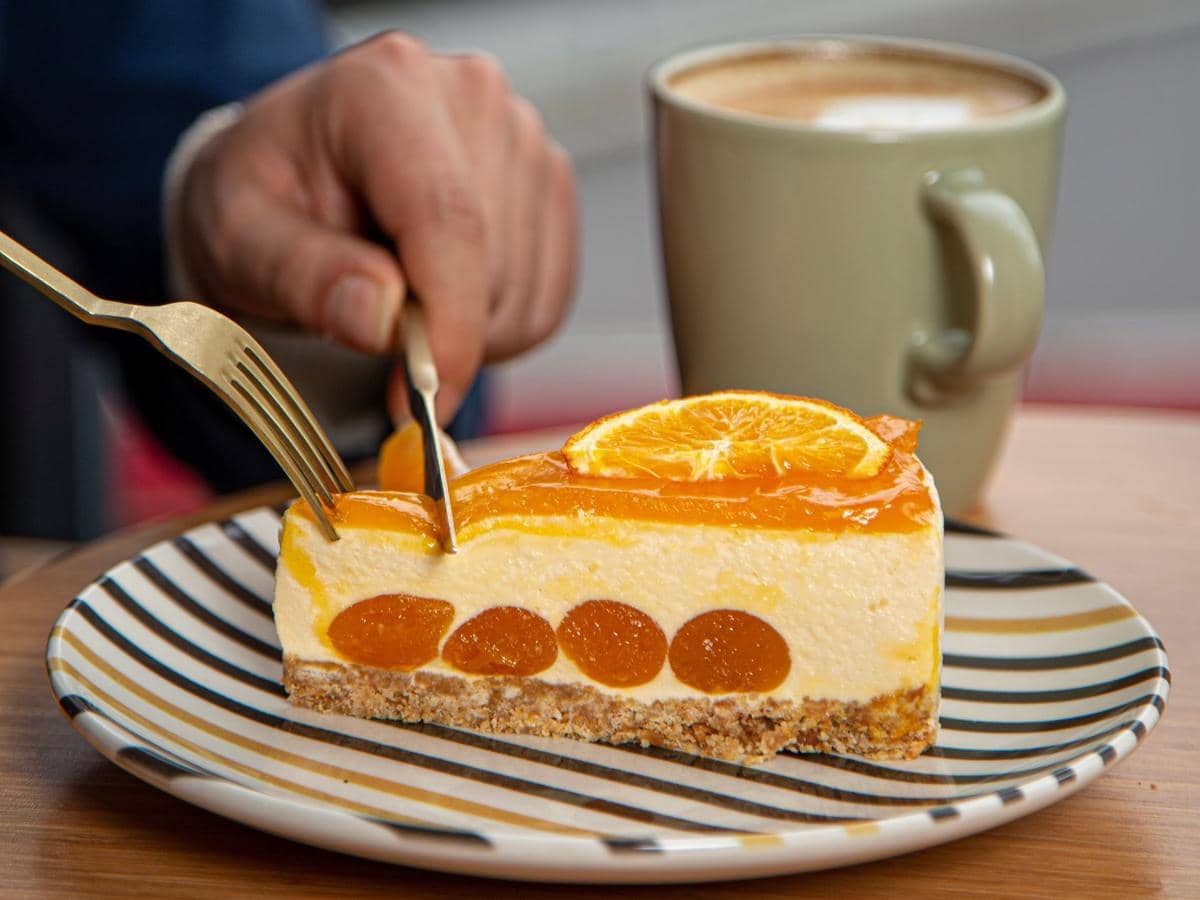 Sepotong orange cheesecake dengan lapisan jeruk segar di atas piring bergaris, disajikan bersama secangkir kopi hangat.