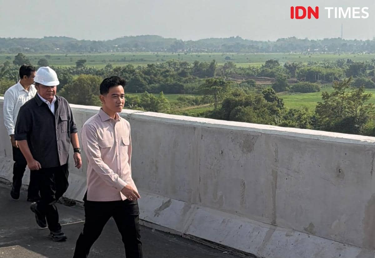 Seorang pria berkemaja muda berjalan di atas jembatan beton bersama rombongan saat meninjau proyek pembangunan di area hijau terbuka.