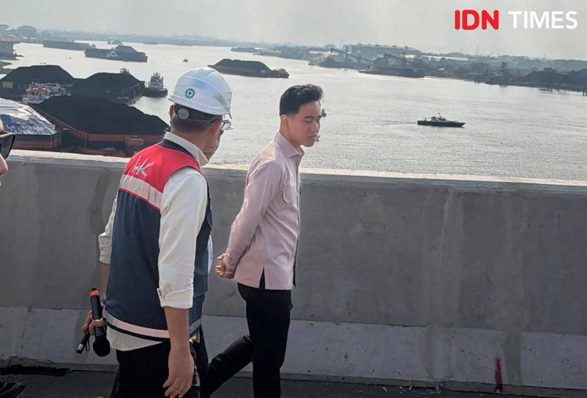 Seorang pria berjas dan seorang pekerja konstruksi berjalan di area pembangunan jembatan di atas Sungai Musi dengan latar kapal dan tongkang.