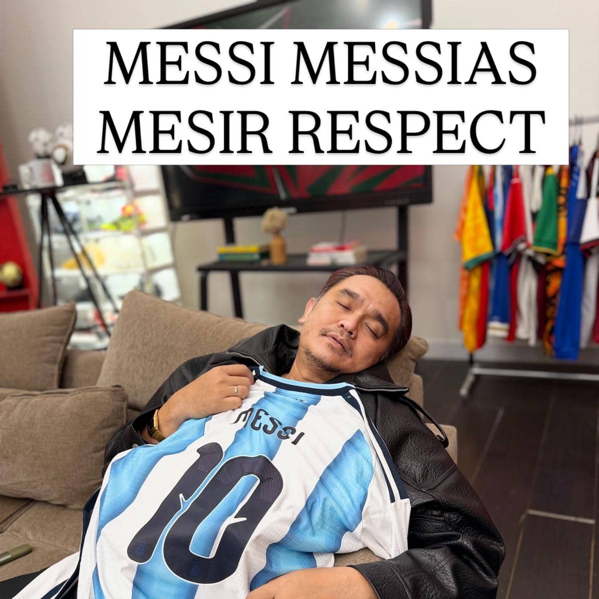 Seorang pria berpose di sofa sambil memeluk jersey bernomor 10 bertuliskan Messi, dengan latar ruangan berisi koleksi pakaian dan televisi.