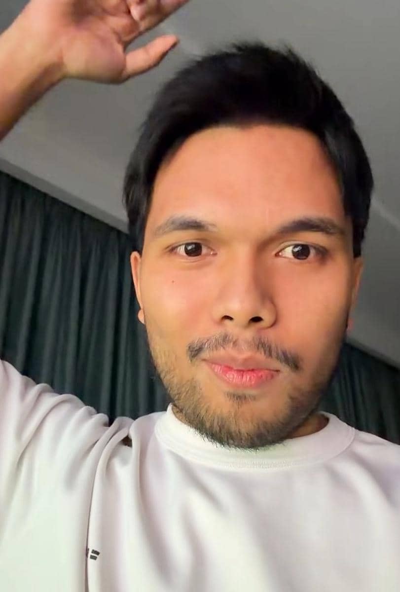 Seorang pria mengenakan kaus putih tersenyum sambil mengangkat tangan di dalam ruangan dengan tirai gelap di belakangnya.