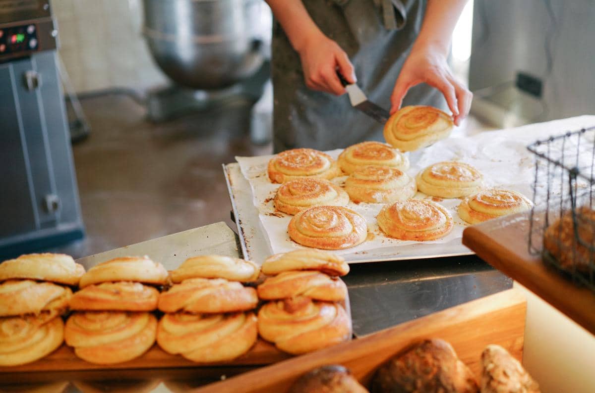 Seorang pembuat roti mengenakan celemek sedang menata kue kering berbentuk spiral di atas loyang di dapur bakery.