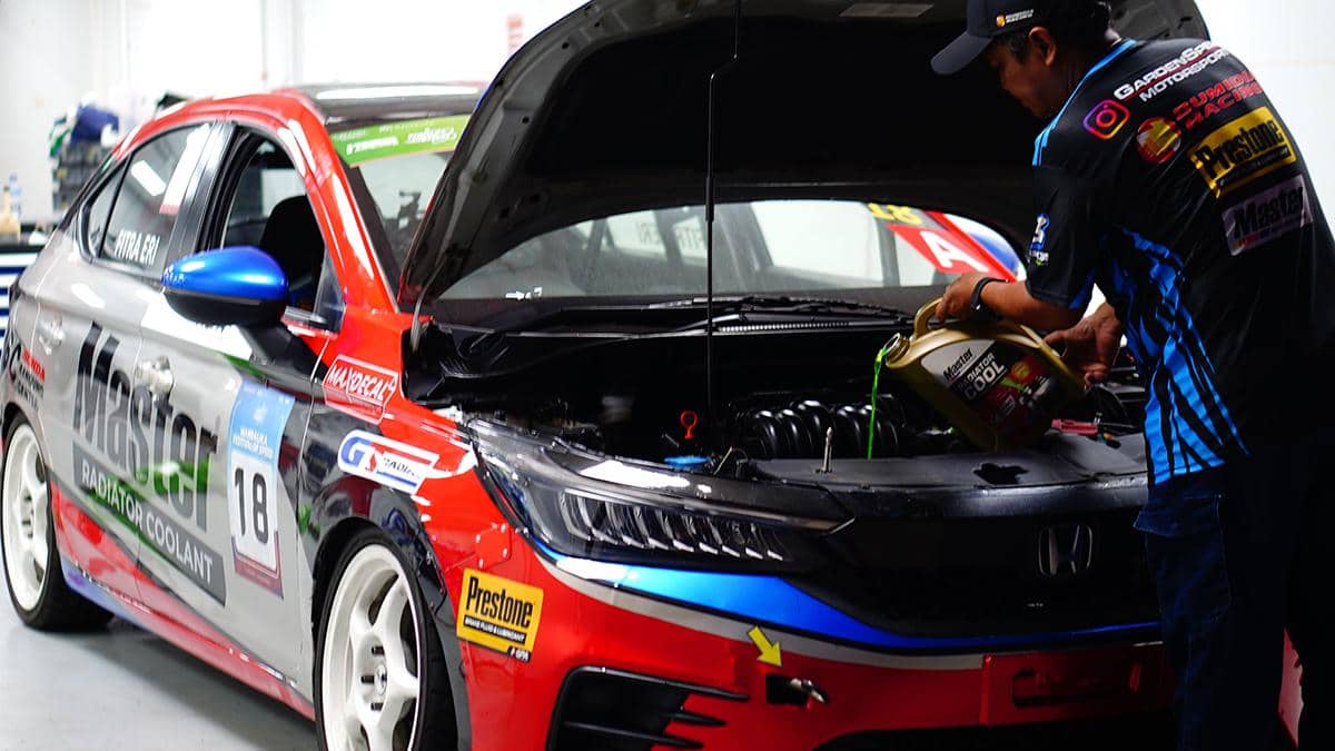 Seorang mekanik menuangkan cairan radiator ke mobil balap Honda berwarna merah, biru, dan abu-abu di area bengkel.
