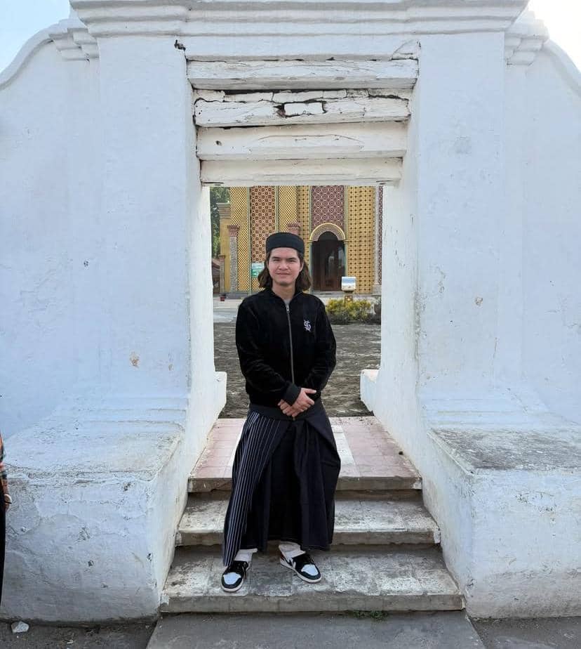 Seorang pria mengenakan jaket hitam dan sarung berdiri di antara gerbang putih tua dengan latar bangunan bercorak khas di Tuban.