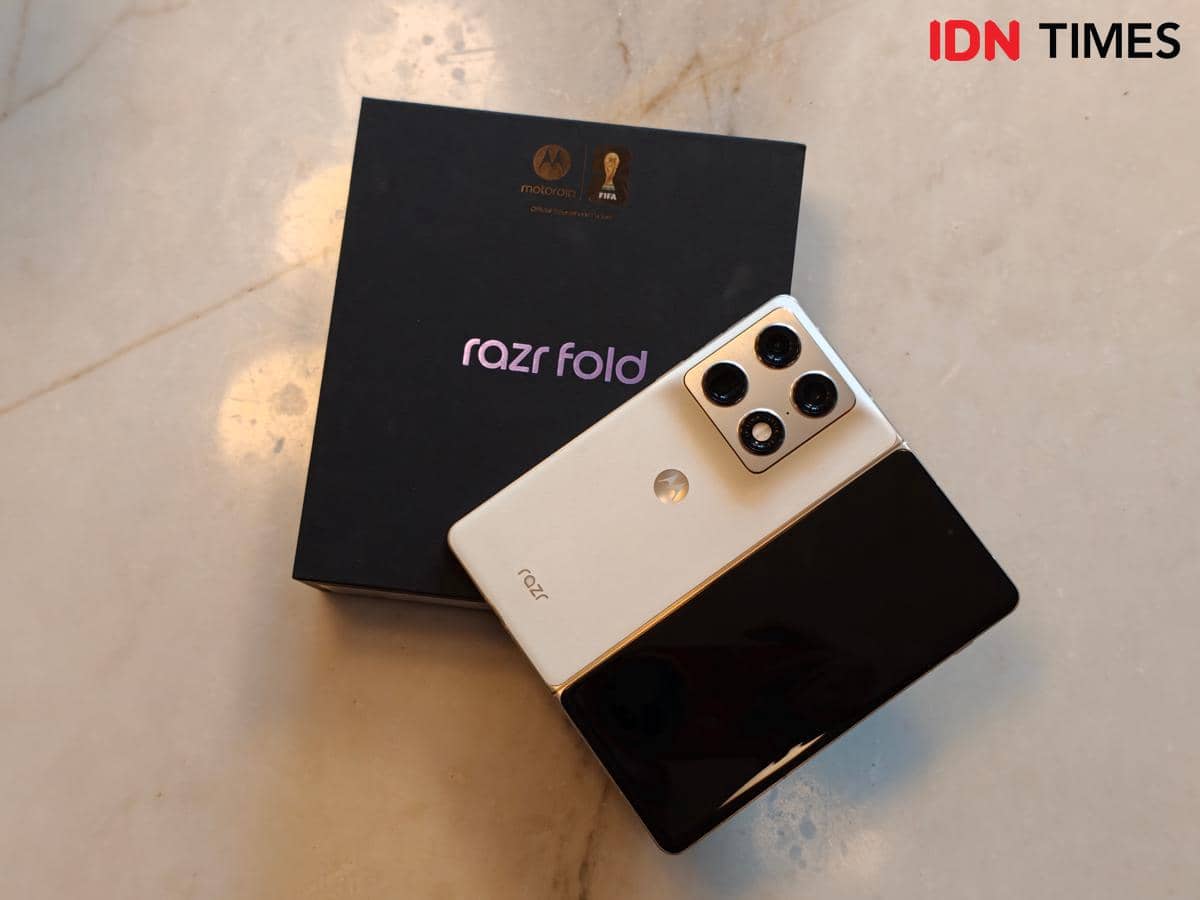 Smartphone lipat Motorola Razr Fold berwarna putih dan hitam dengan tiga kamera belakang, diletakkan di atas kotak kemasan hitam.