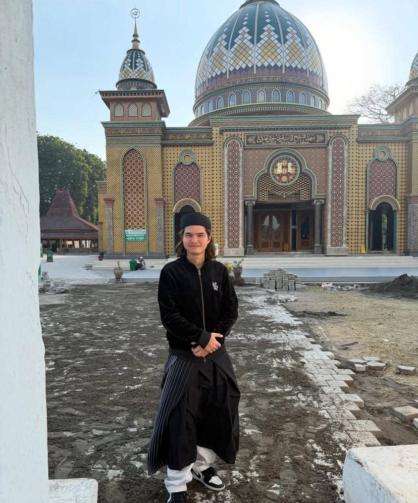 Seorang pria mengenakan pakaian hitam dan sarung berdiri di depan masjid megah dengan kubah besar berwarna emas dan biru di Tuban.