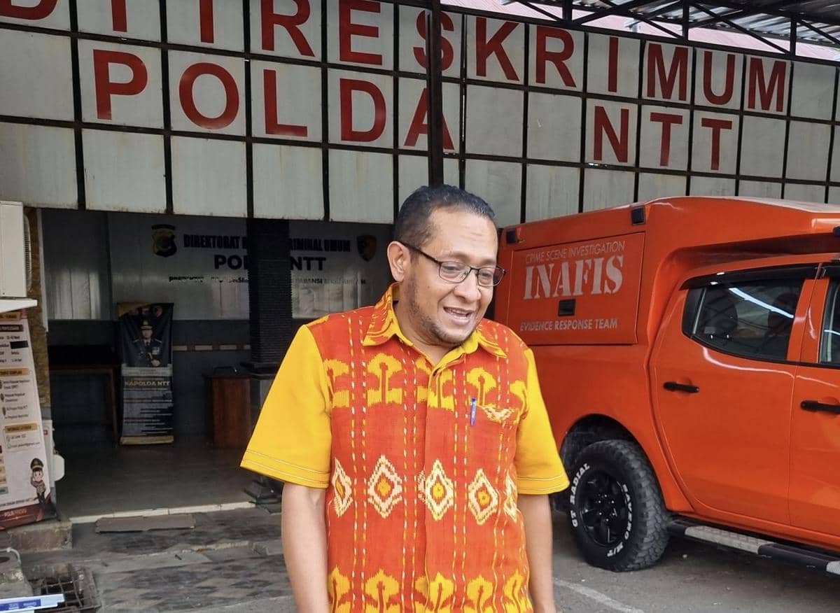 Seorang pria mengenakan kemeja motif tenun merah dan kuning berdiri di depan gedung Ditreskrimum Polda NTT dengan mobil INAFIS di sampingnya.