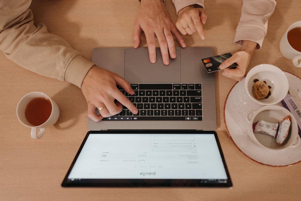 Dua orang menggunakan laptop sambil memegang kartu kredit di meja dengan dua cangkir teh dan piring berisi camilan.