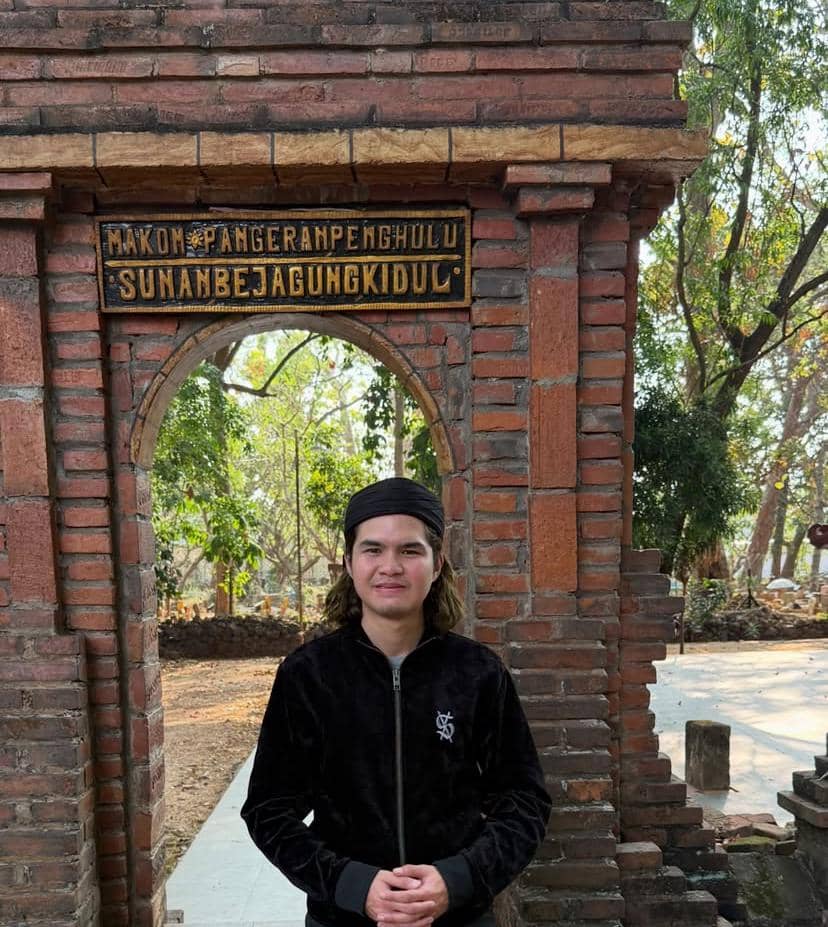 Seorang pria mengenakan pakaian hitam dan ikat kepala berdiri di depan gerbang bata bertuliskan Makom Pangeran Penghulu Sunan Bejagung Kidul di Tuban.