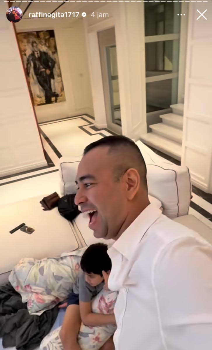 Seorang pria tersenyum lebar di ruang tamu modern berwarna putih, sementara seorang anak duduk di sofa dengan selimut bermotif bunga.