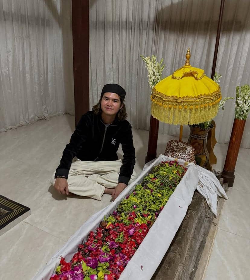 Seorang pria duduk bersila di samping makam yang dihiasi bunga warna-warni dan payung kuning di ruangan tertutup.