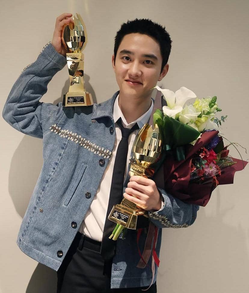 Seorang pria mengenakan jaket denim dan dasi hitam memegang dua piala emas serta buket bunga sambil tersenyum ke arah kamera.