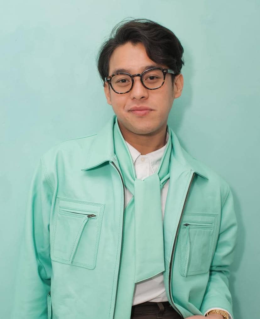 Seorang pria berkacamata mengenakan jaket dan syal berwarna hijau mint dengan latar dinding berwarna senada.