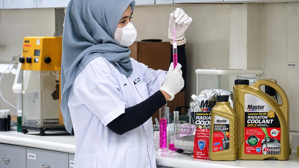 Seorang peneliti perempuan berhijab mengenakan jas laboratorium dan masker sedang menguji cairan berwarna merah muda di laboratorium kimia.