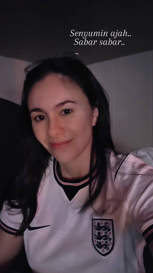 Seorang perempuan mengenakan jersey tim nasional Inggris tersenyum tipis dengan tulisan motivasi di latar bertuliskan 'Senyumin ajah.. Sabar sabar..'.