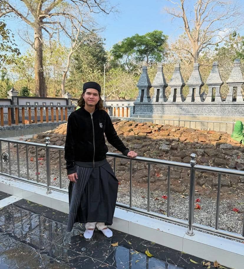 Seorang pria mengenakan pakaian hitam dan sarung berdiri di area wisata religi Tuban dengan latar makam dan pepohonan rindang.