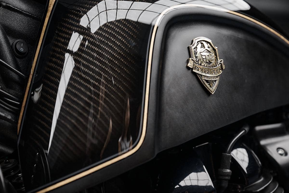 Tangki motor custom Rough Crafts berwarna hitam dengan detail serat karbon dan emblem logam berlogo singa di sisi bodi.